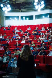 Salle de conférence avec des gens dans le public et une personne qui parle au micro de dos.