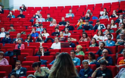 Salle de conférence avec des gens dans le public et une personne qui parle au micro de dos.